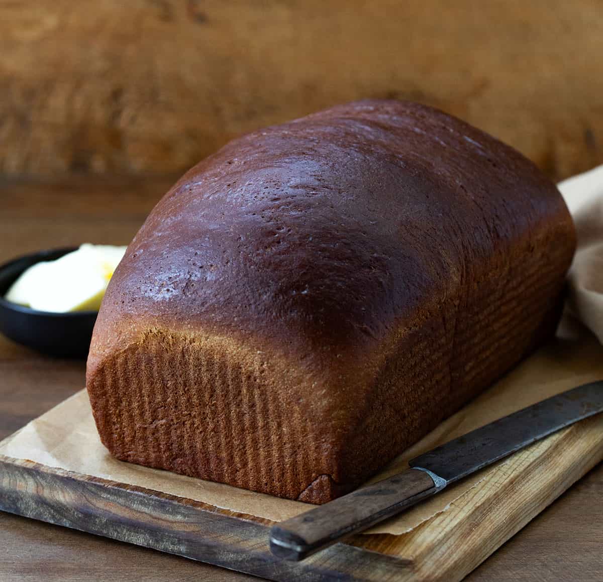 This homemade Pumpernickel Bread is a rye bread that has a rich, slightly sweet flavor with a dense, hearty texture. It's also perfect for sandwiches or a bowl of soup or stew. Whole Pumpernickel Bread loaf on a cutting board on a wooden table.