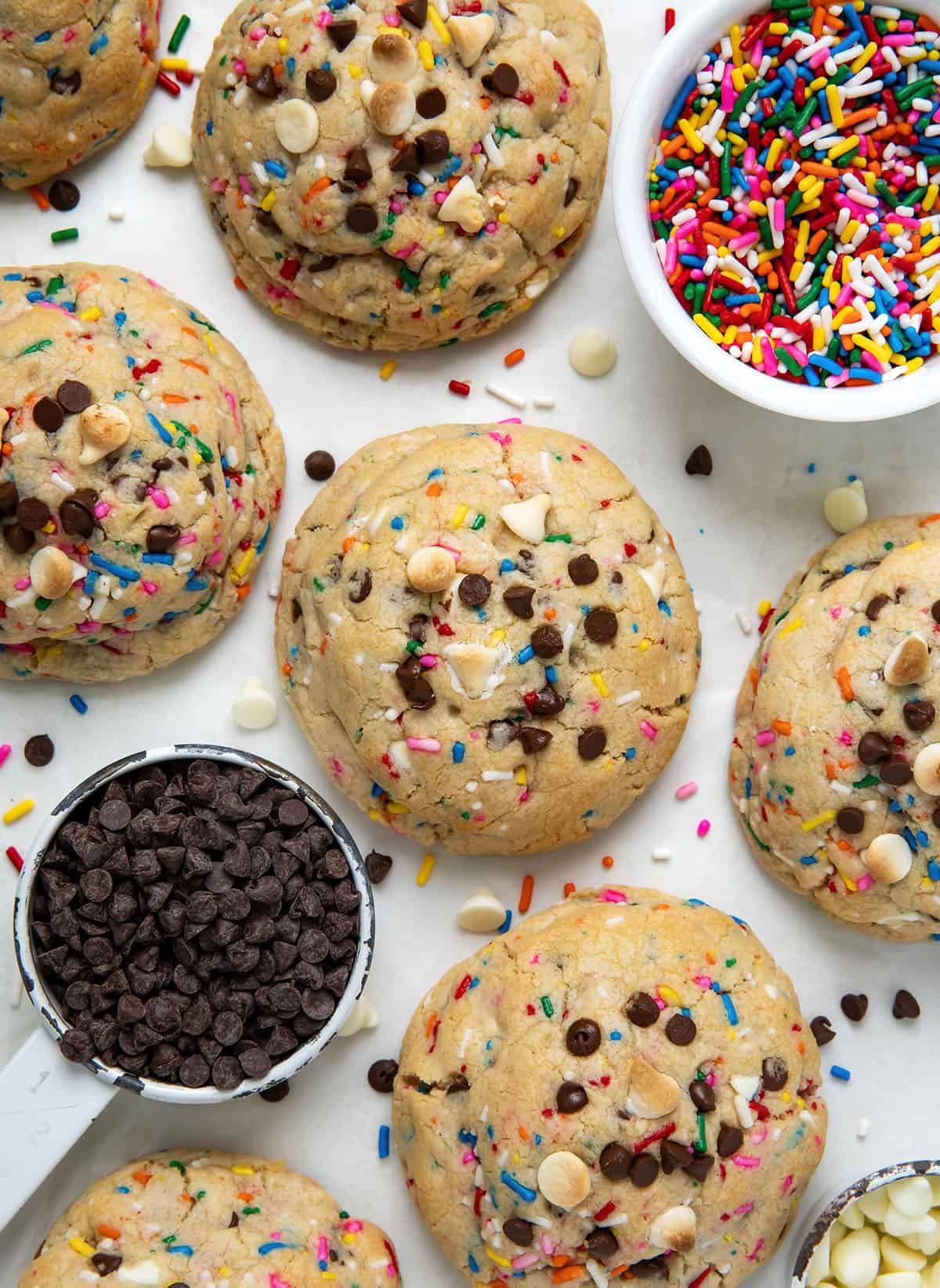 Thick and Chewy Sprinkles Cookies on a white table with sprinkles and mini chocolate chips and white chocolate chips from overhead.