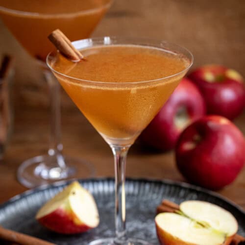 Two Apple Cider Martini's on a wooden table with apples and cinnamon stick.