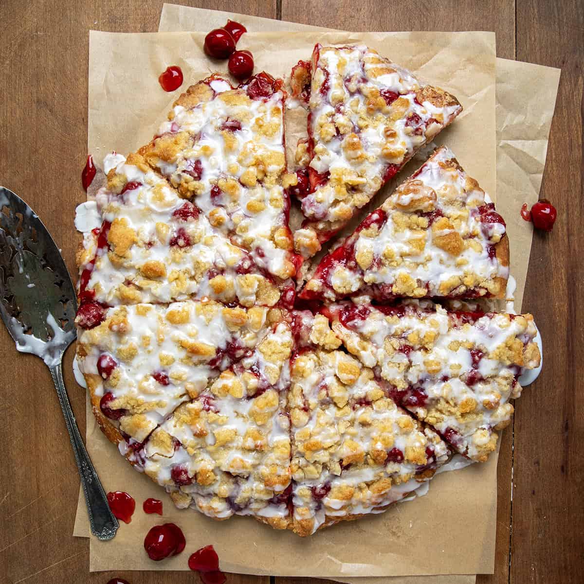 Whole Cherry Dessert Pizza cut into pieces on parchment paper on a wooden table with a cake server and cherries around.