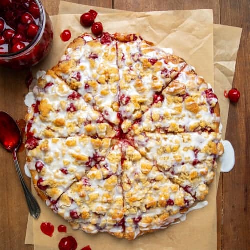 Whole Cherry Dessert Pizza on parchment paper on a wooden table with a jar of cherries from overhead.