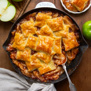 Skillet of Apple Pie Puff Pastry Bake with a spoon in it on a wooden table from overhead.