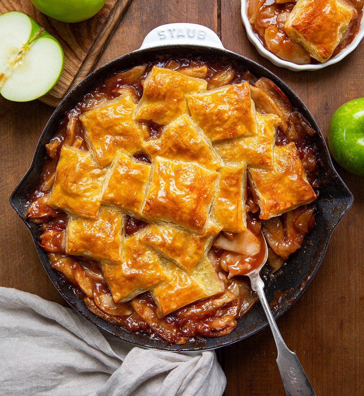 Skillet of Apple Pie Puff Pastry Bake with a spoon in it on a wooden table from overhead. 