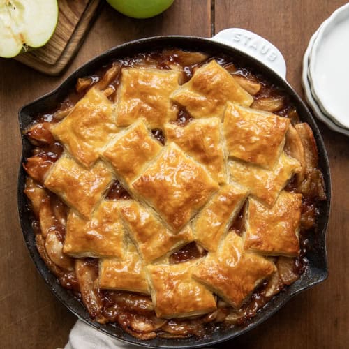 White Skillet of Apple Pie Puff Pastry Bake showing golden puffy pastry on a wooden table with apples from overhead.