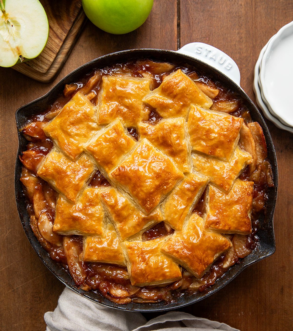 White Skillet of Apple Pie Puff Pastry Bake showing golden puffy pastry on a wooden table with apples from overhead.