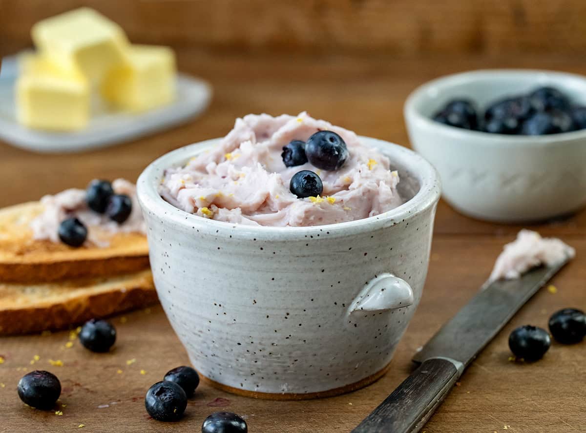 Small jar of Lemon Blueberry Butter on a wooden table with butter, blueberries, and a knife.