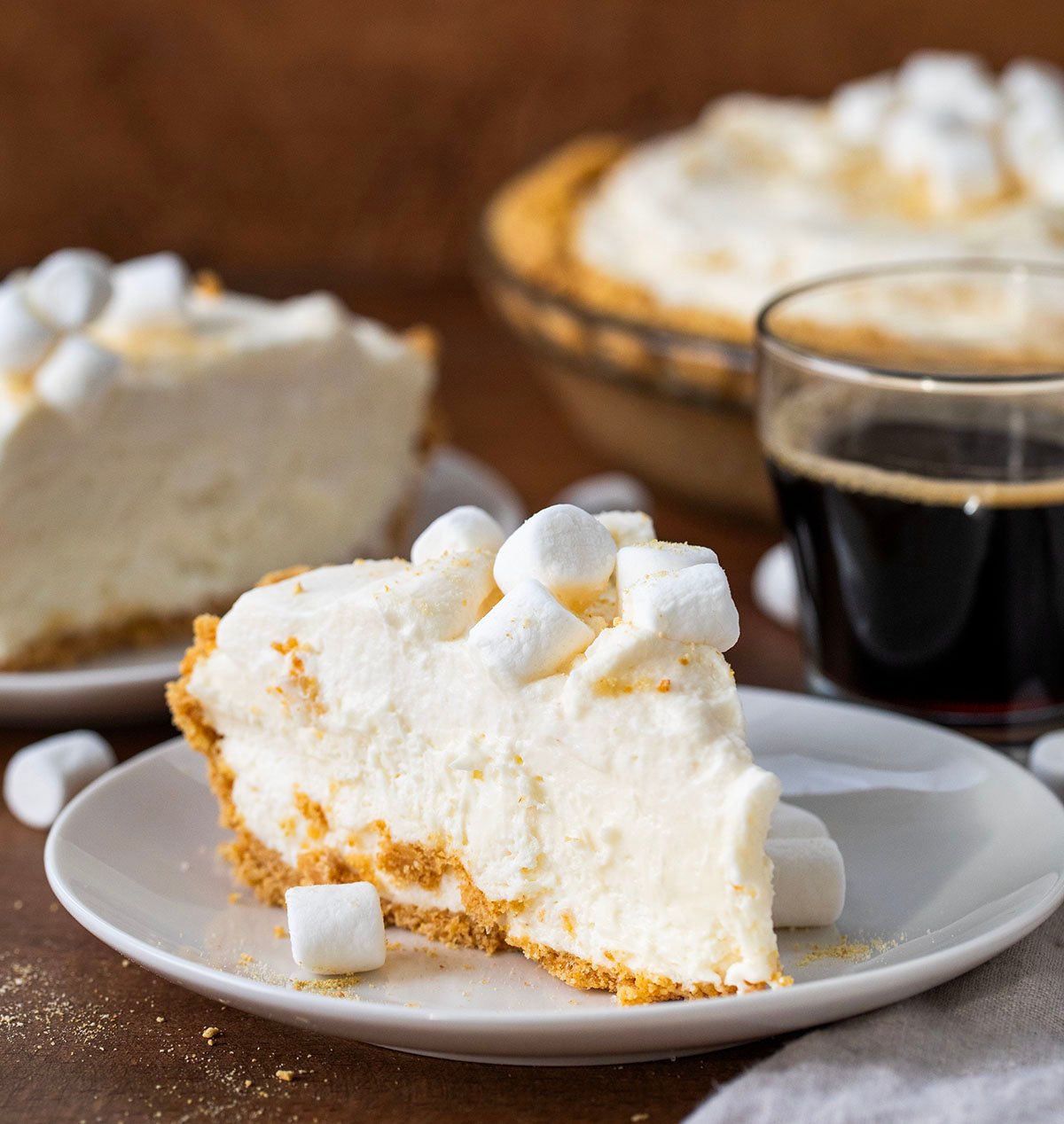 White plates with Marshmallow Cream Cheesecake on them with mini marshmallows and coffee on a wooden table.