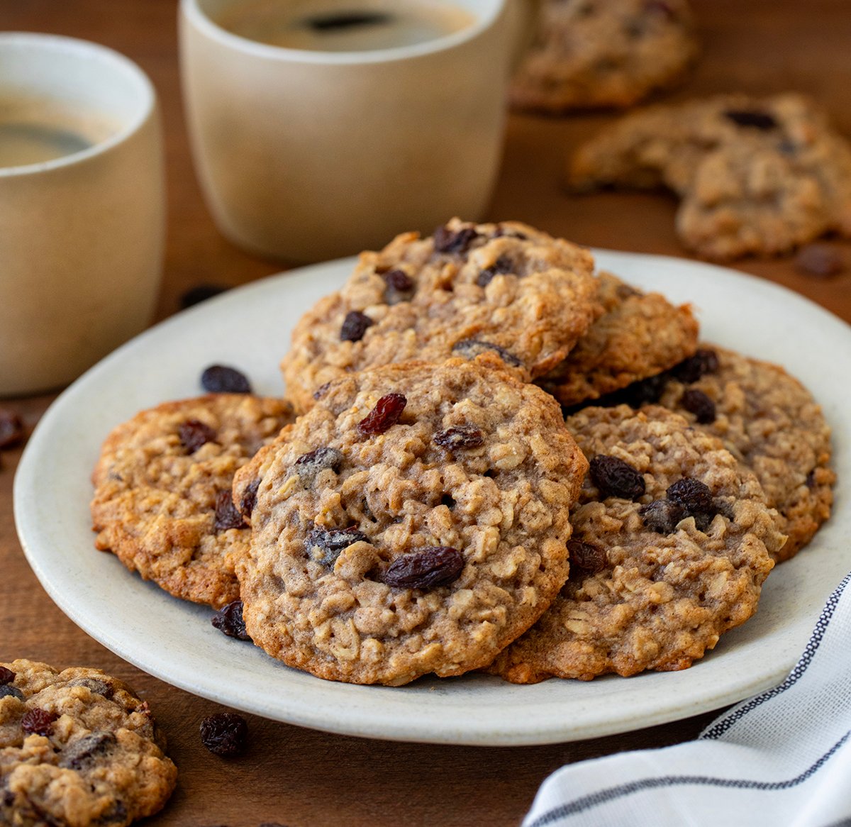 Plate of Oatmeal Raisin Cookies on a wooden table with coffee cups.