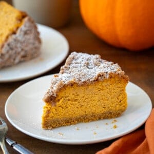 Slice of Pumpkin Crumble Cheesecake on a white plate with a pumpkin in the background.