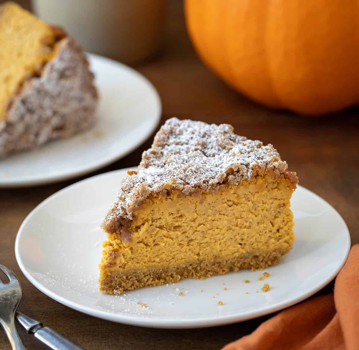 Slice of Pumpkin Crumble Cheesecake on a white plate with a pumpkin in the background.