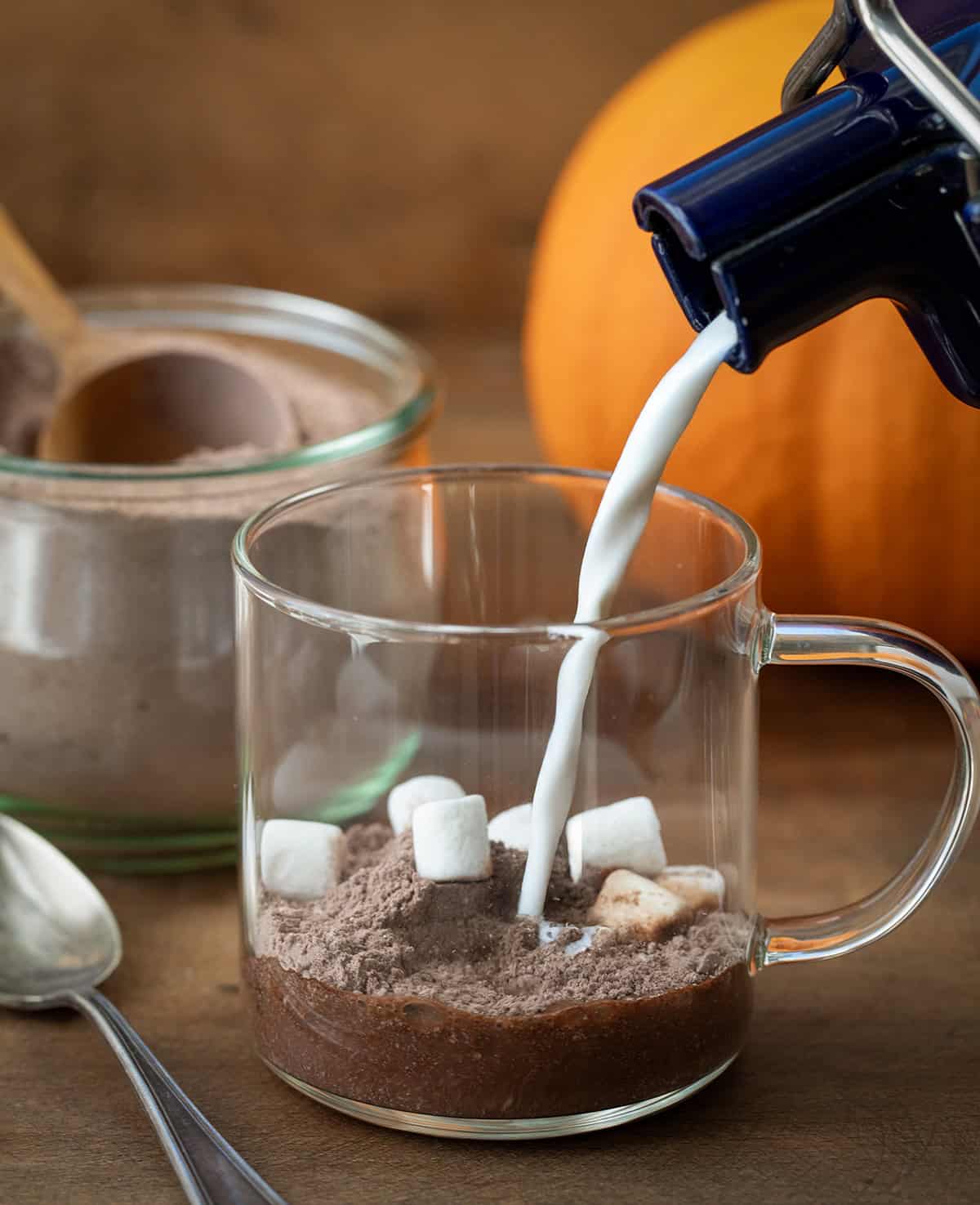 Pouring milk into a glass mug with Pumpkin Spice Hot Cocoa Mix in it and mini marshmallows.