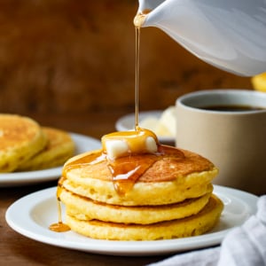 Pouring syrup over stack of Cornmeal Pancakes with butter.