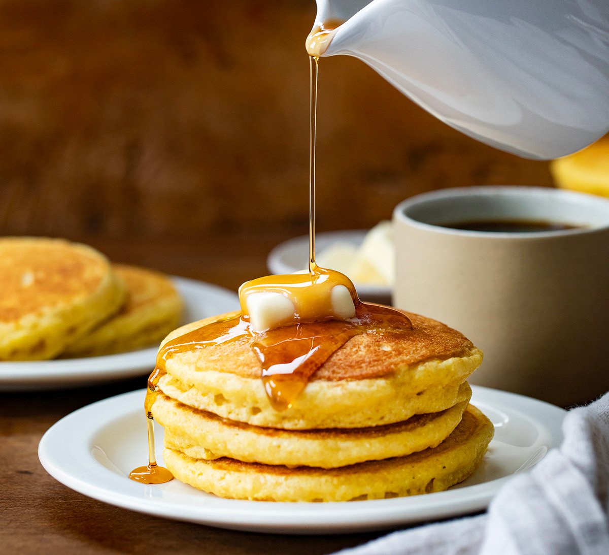 Pouring syrup over stack of Cornmeal Pancakes with butter.