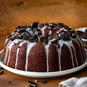 Oreo Bundt Cake on a white cake plate on a wooden table with oreos around.