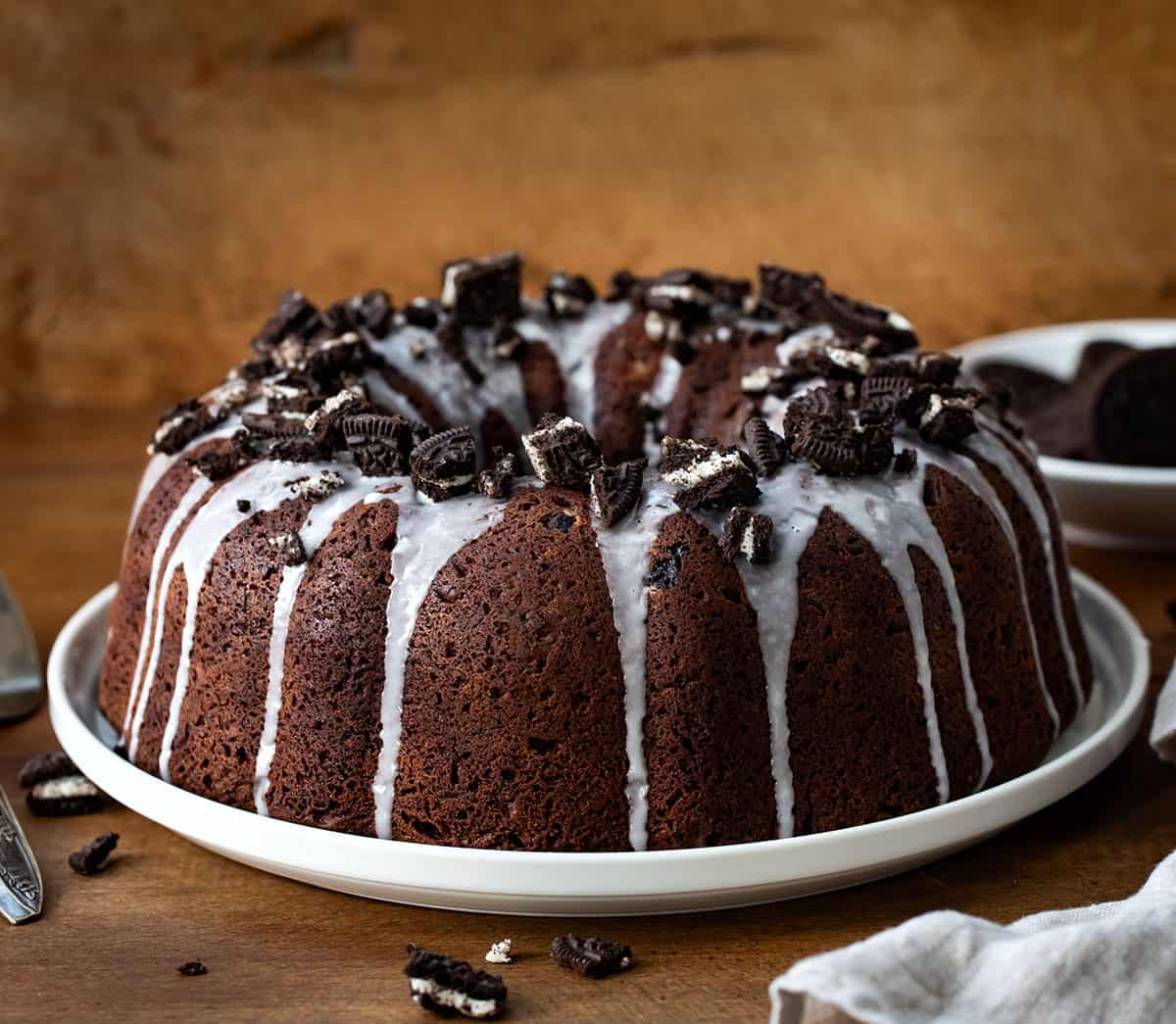 Oreo Bundt Cake on a white cake plate on a wooden table with oreos around.