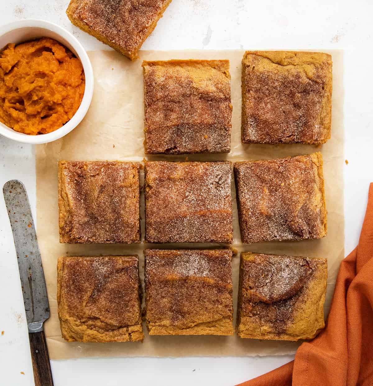 Pumpkin Snickerdoodle Brownies on a white table with pumpkin puree, an orange towel, and a knife. 