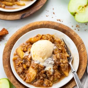 Plates of Slow Cooker Apple Crisp on a white table with cinnamon sticks and apples.