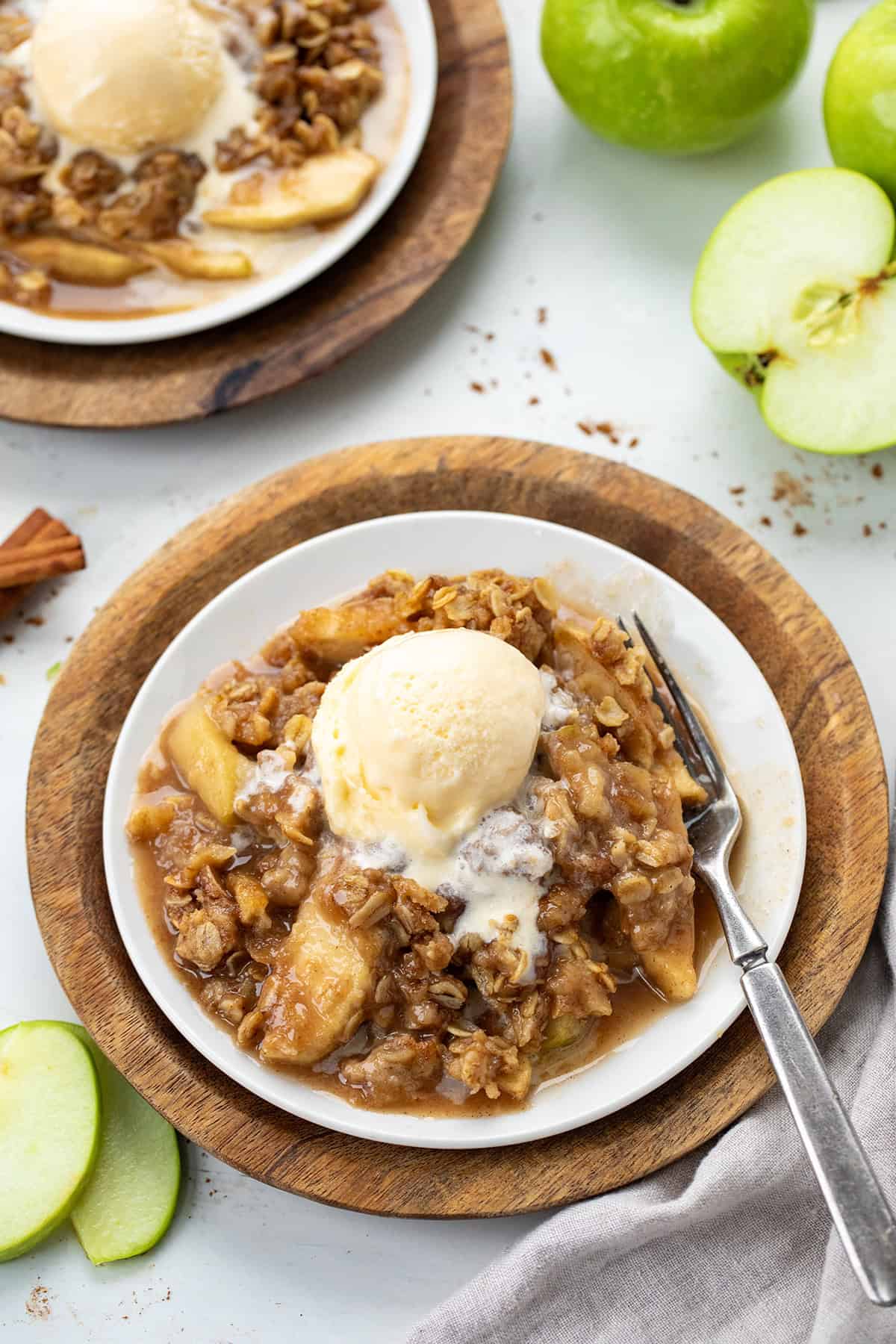 Plates of Slow Cooker Apple Crisp on a white table with cinnamon sticks and apples.