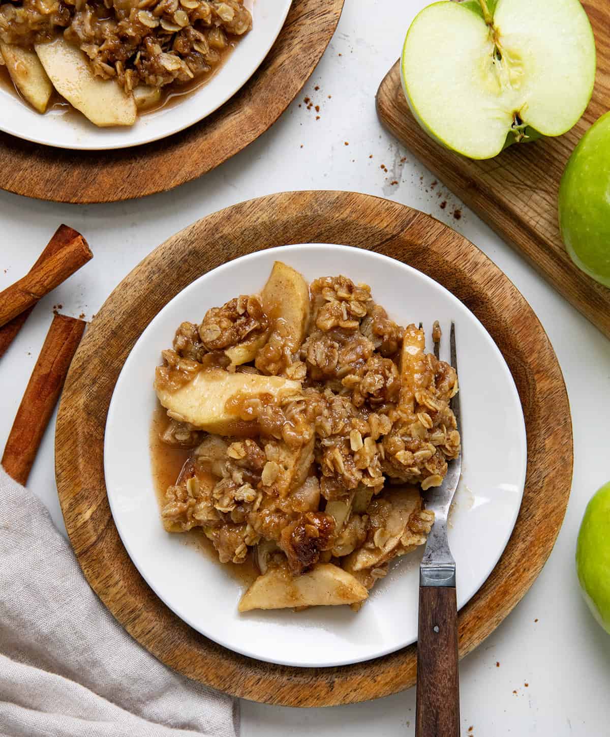 Plates of Slow Cooker Apple Crisp on a white table with cinnamon sticks and apples.