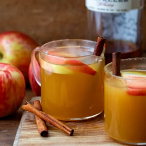 Glasses of Spiked Apple Cider on a wooden cutting board with fresh apples and cinnamon sticks.