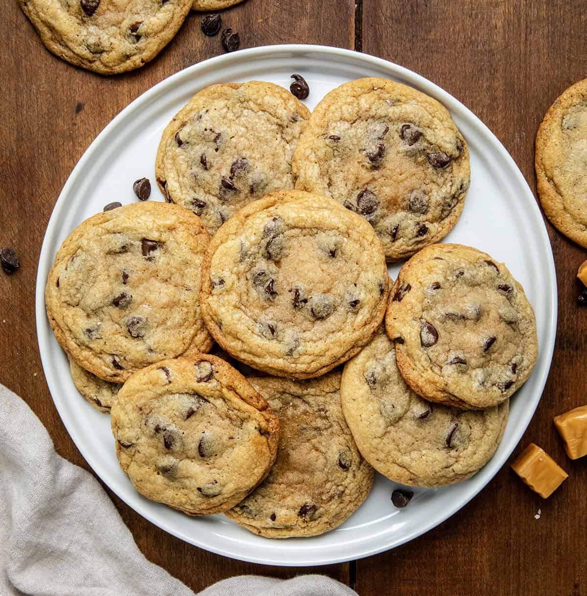 White plate of Caramel Stuffed Chocolate Chip Cookies on a wooden table with caramels and chocolate chips from overhead.