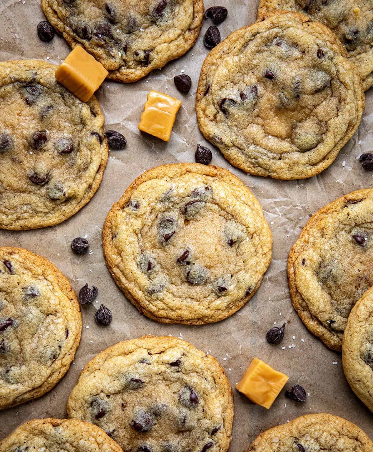 Caramel Stuffed Chocolate Chip Cookies on brown parchment paper with caramels and chocolate chips from overhead.