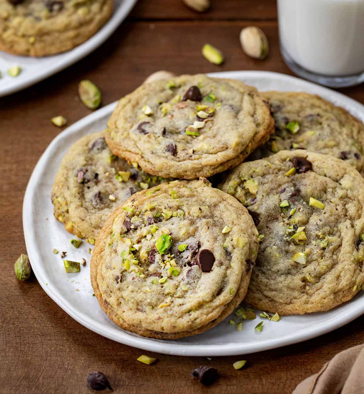 Pistachio Chocolate Chip Cookies on a white plate on a wooden table with milk in the background.