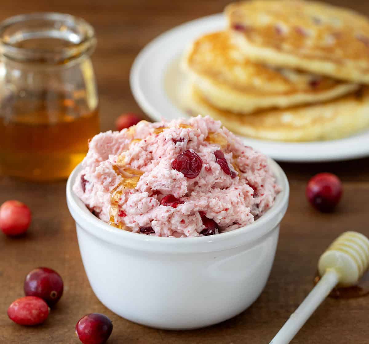 Bowl of Cranberry Honey Butter on a wooden table with pancakes and honey in background.