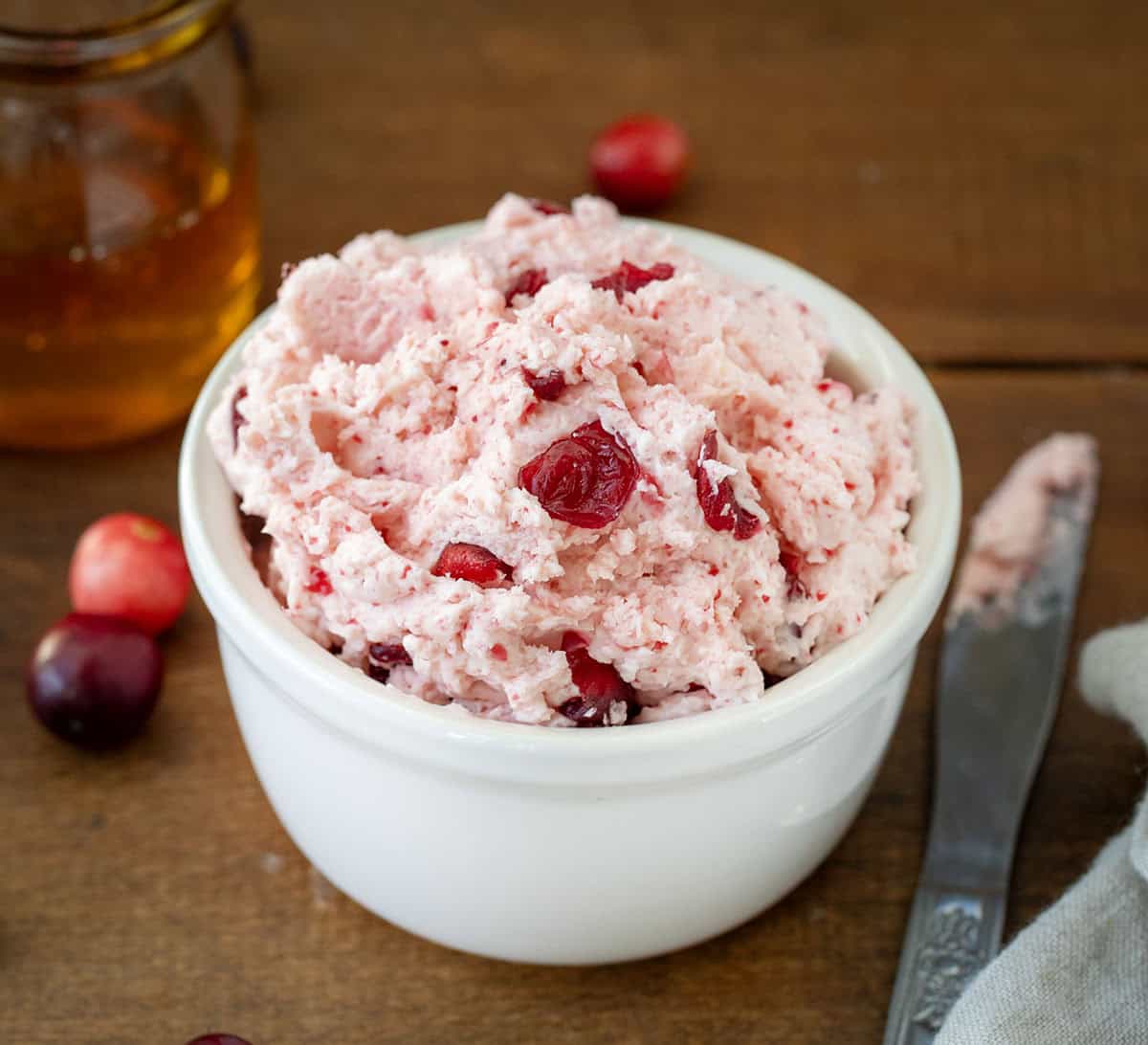 Bowl of Cranberry Honey Butter on a wooden table with knife and honey.