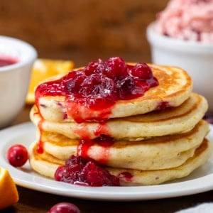 Stack of Cranberry Orange Buttermilk Pancakes on a white plate with cranberry sauce on a wooden table.