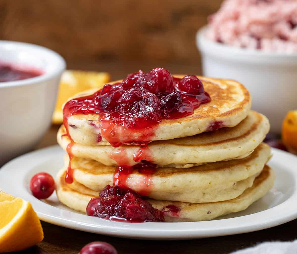 Stack of Cranberry Orange Buttermilk Pancakes on a white plate with cranberry sauce on a wooden table. 