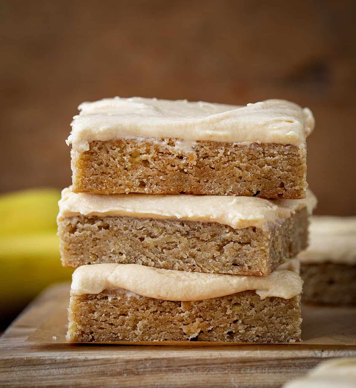 Stack of Browned Butter Banana Bars With Salted Caramel Buttercream on a wooden table close up with bananas in the background.