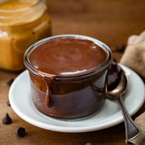 Jar of Peanut Butter Hot Fudge on a plate with peanut butter in the background.