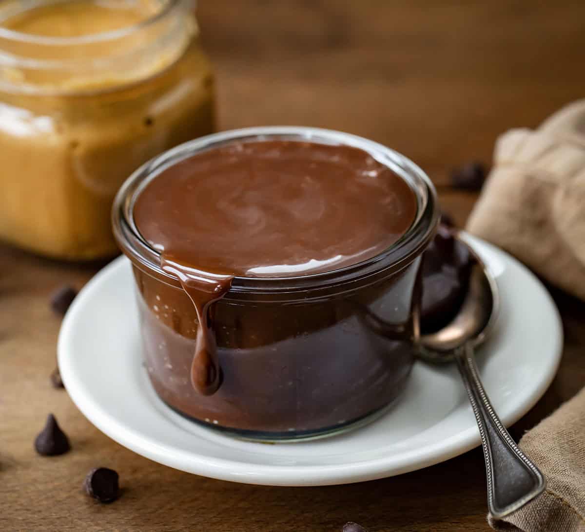 Jar of Peanut Butter Hot Fudge on a plate with peanut butter in the background.