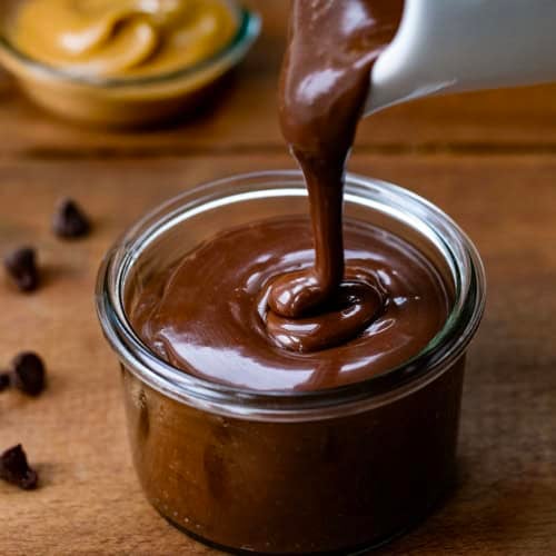 Pouring Peanut Butter Hot Fudge into a jar on a wooden table with creamy peanut butter.