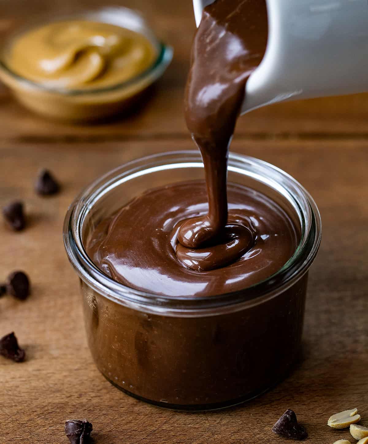 Pouring Peanut Butter Hot Fudge into a jar on a wooden table with creamy peanut butter.