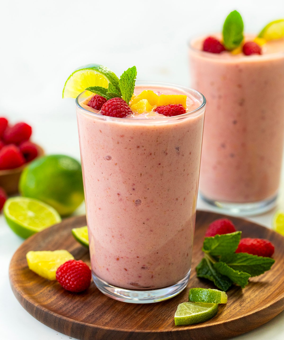 Tropical Smoothies on a white table with fresh limes, raspberries, mango, and banana around. 