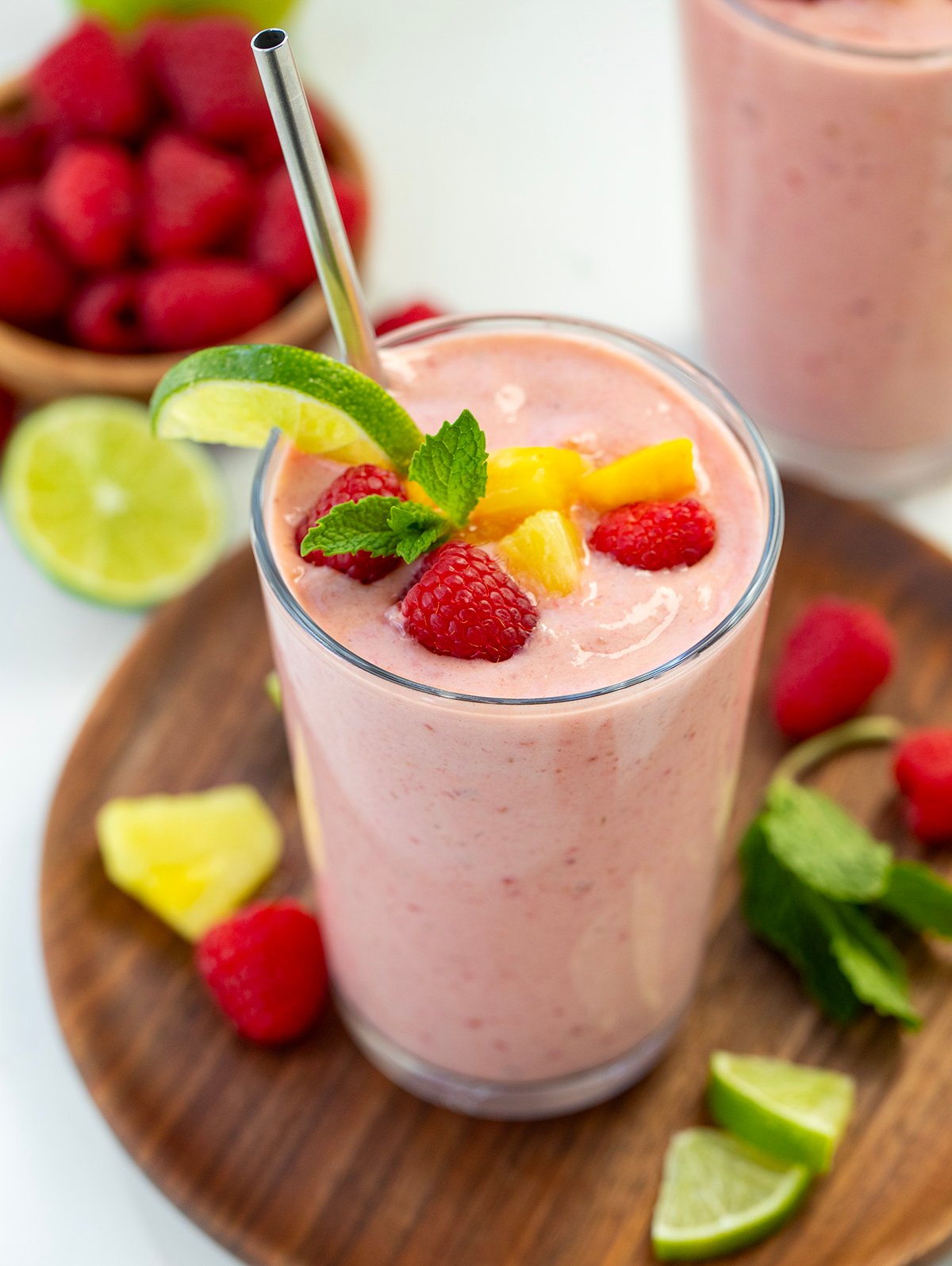 Tropical Smoothies on a white table with fresh limes, raspberries, mango, and banana around.