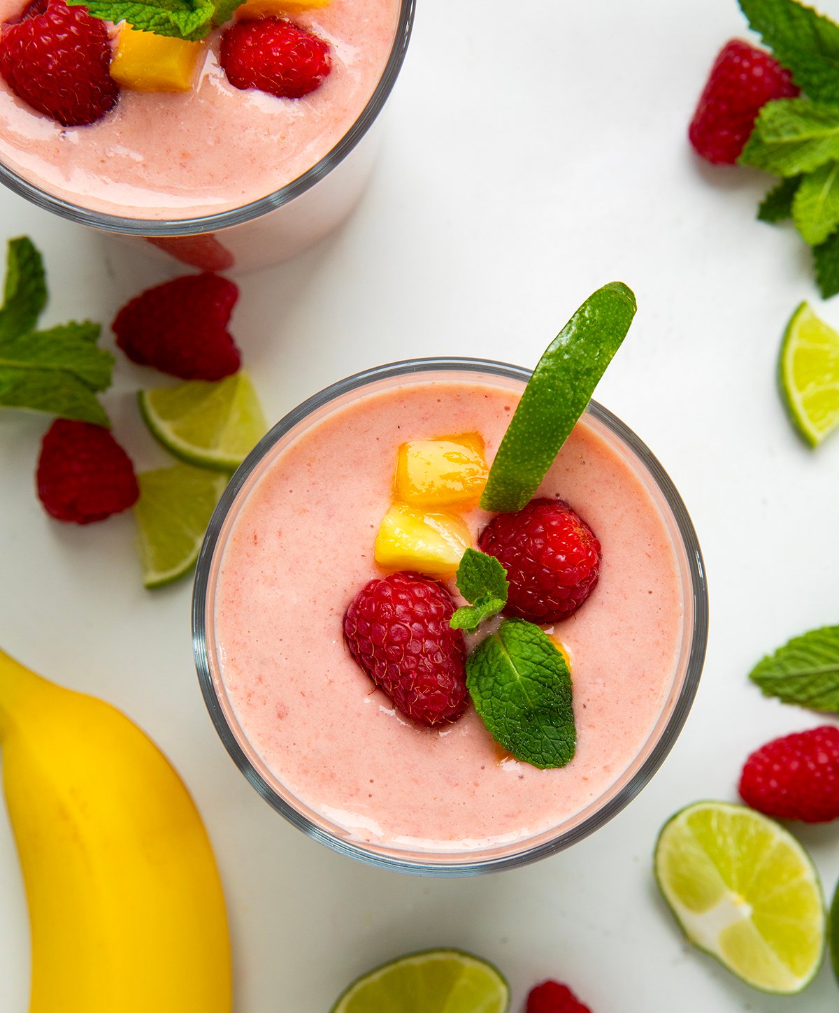 Tropical Smoothies on a white table with fresh limes, raspberries, mango, and banana around from overhead.