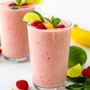 Tropical Smoothies on a white table with fresh limes, raspberries, mango, and banana around.