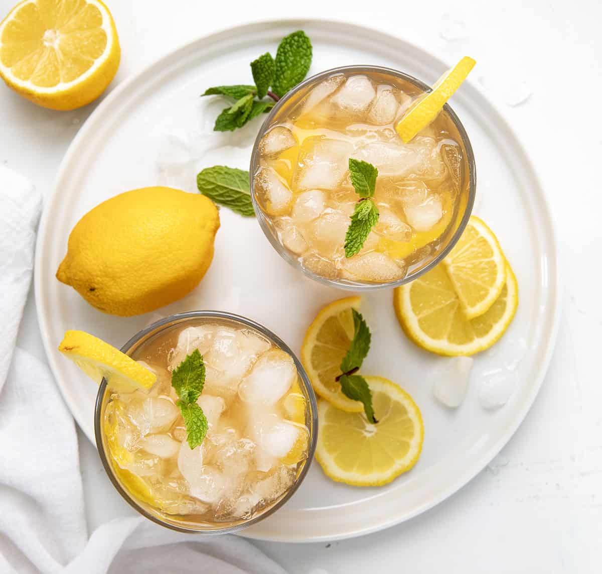 Glasses of Arnold Palmer on a white tray with fresh lemon, ice, and mint leaves from overhead.