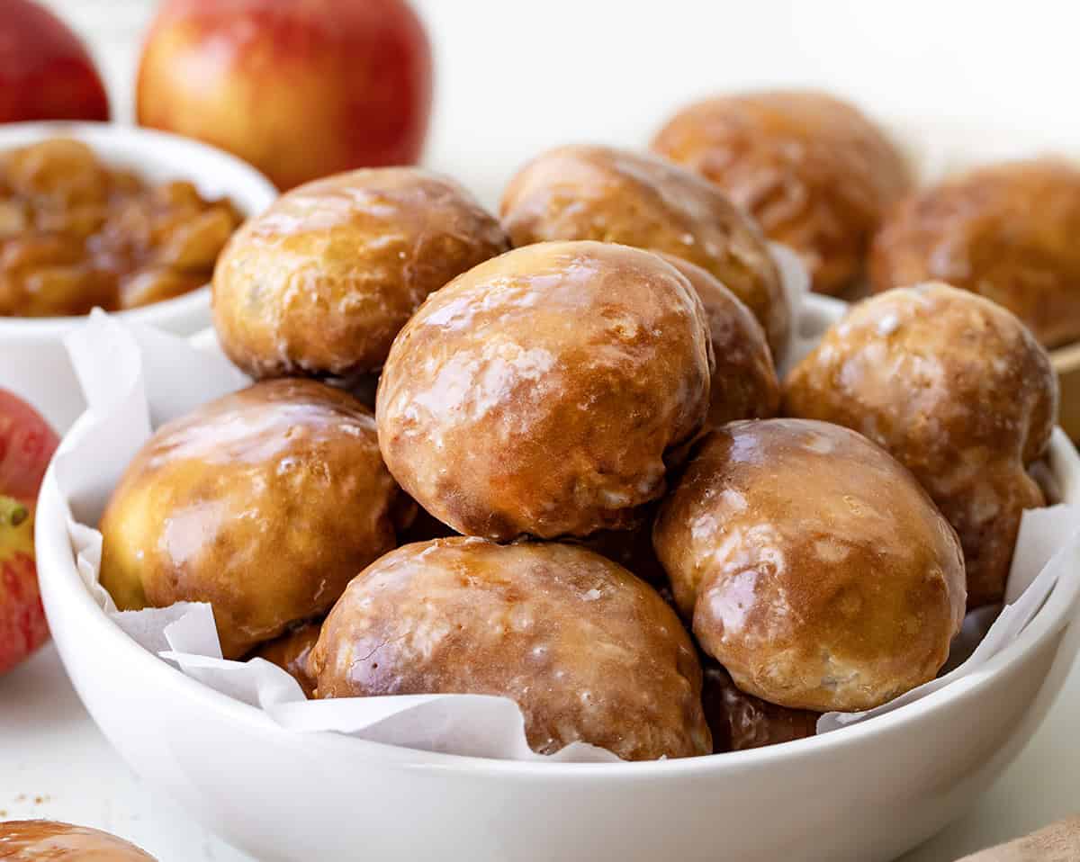 These easy Apple Fritter Bombs are made with biscuit dough, apple pie filling, and a sweet glaze, just like a mini apple fritter! Close up of Apple Fritter Bombs in a white bowl with apples in the background.
