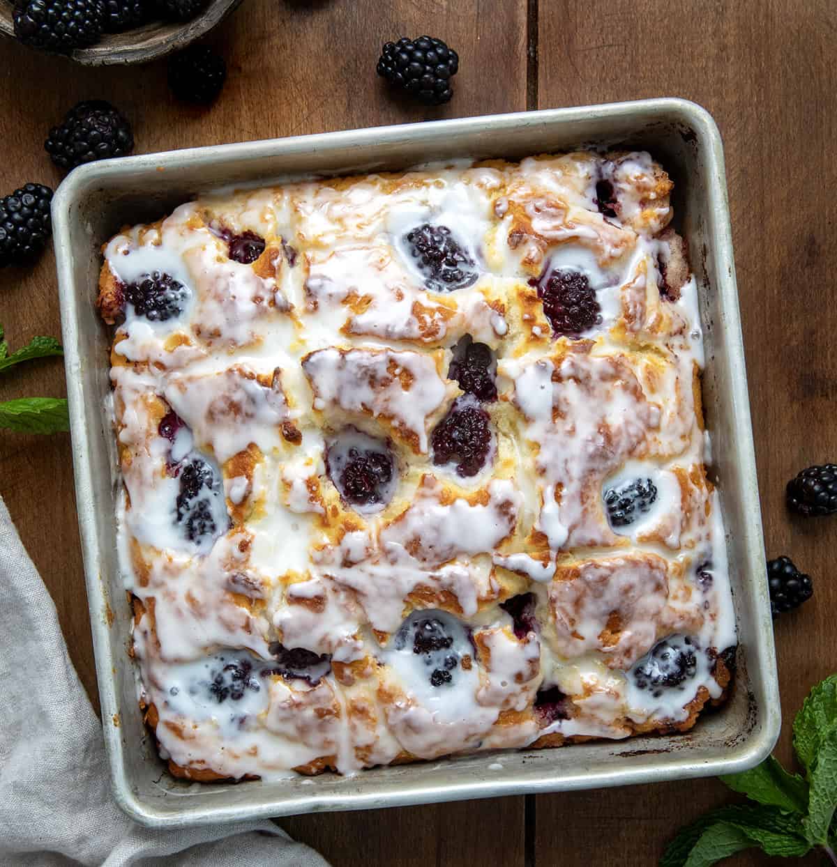 Blackberry Butter Swim Biscuits are buttery buttermilk biscuits filled with fresh blackberries and baked in melted butter, then topped with a simple, sweet glaze! Pan of Blackberry Butter Swim Biscuits on a wooden table from overhead.