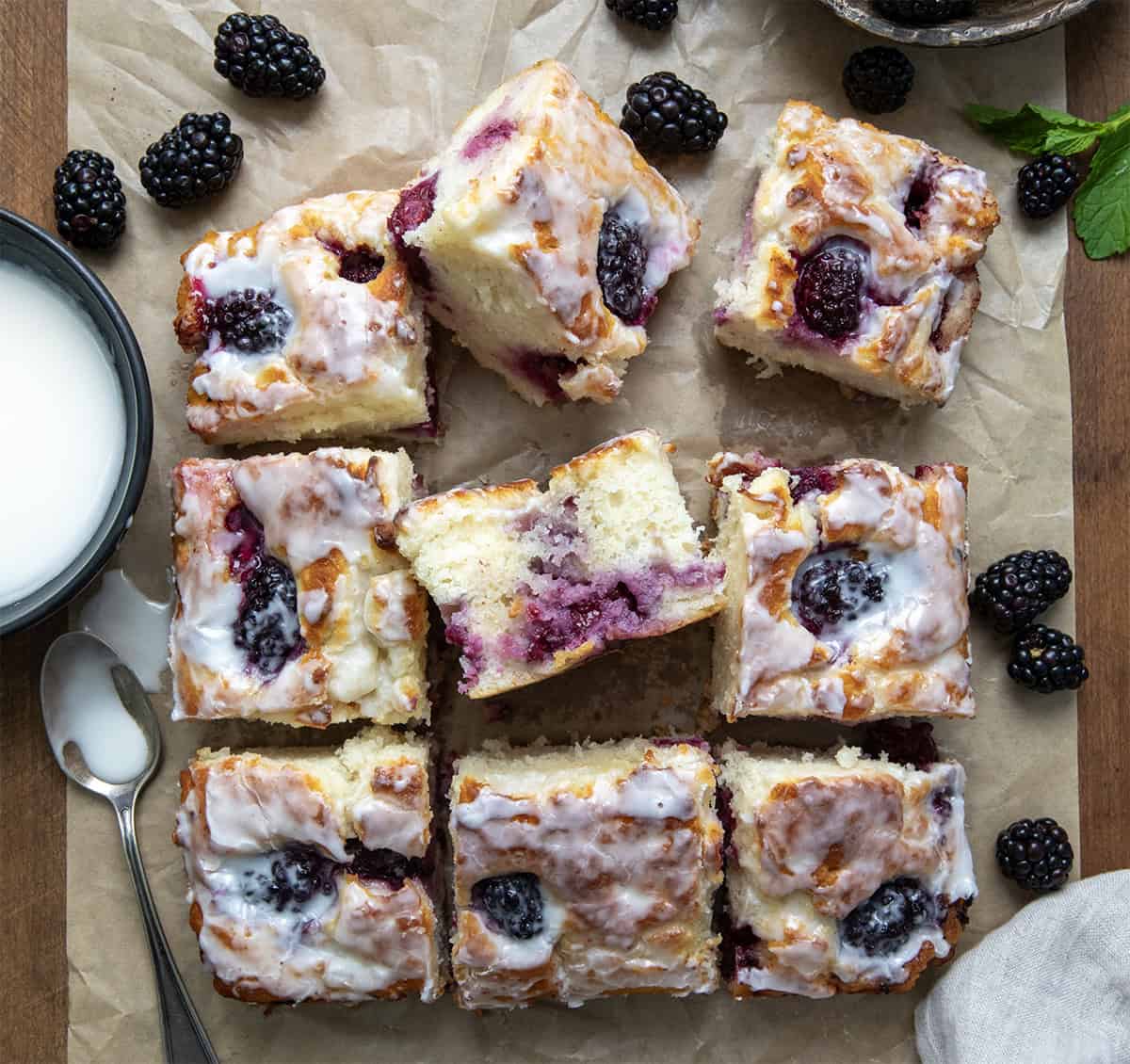 Blackberry Butter Swim Biscuits are buttery buttermilk biscuits filled with fresh blackberries and baked in melted butter, then topped with a simple, sweet glaze! Blackberry Butter Swim Biscuits cut into squares on a piece of brown parchment paper on a wooden table.