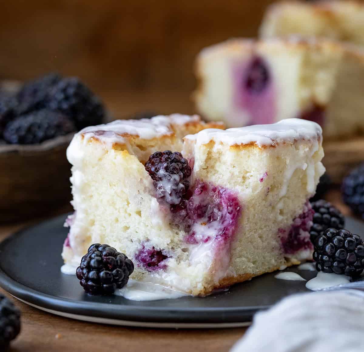 Blackberry Butter Swim Biscuits are buttery buttermilk biscuits filled with fresh blackberries and baked in melted butter, then topped with a simple, sweet glaze! Blackberry Butter Swim Biscuit on a plate on a wooden table.