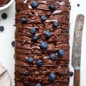 Whole Blueberry Brownie Bread on a white table with fresh blueberries, ganache, and a knife.