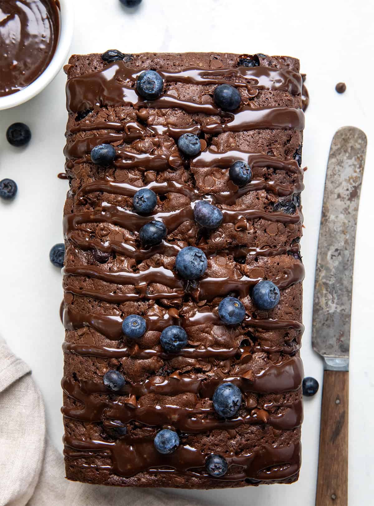 Blueberry Brownie Bread is a rich, fudgy chocolate dessert loaf packed with juicy blueberries and chocolate chips. It’s the perfect mix of a brownie and a quick bread, moist, dense, and full of flavor in every bite! Whole Blueberry Brownie Bread on a white table with fresh blueberries, ganache, and a knife.