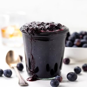 Jar of Bourbon Blueberry Jam with a glass of bourbon behind and fresh blueberries.