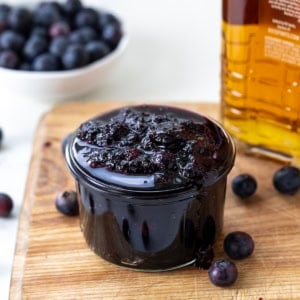 Jar of Bourbon Blueberry Jam on a cutting board on a white table with bourbon and fresh blueberries.