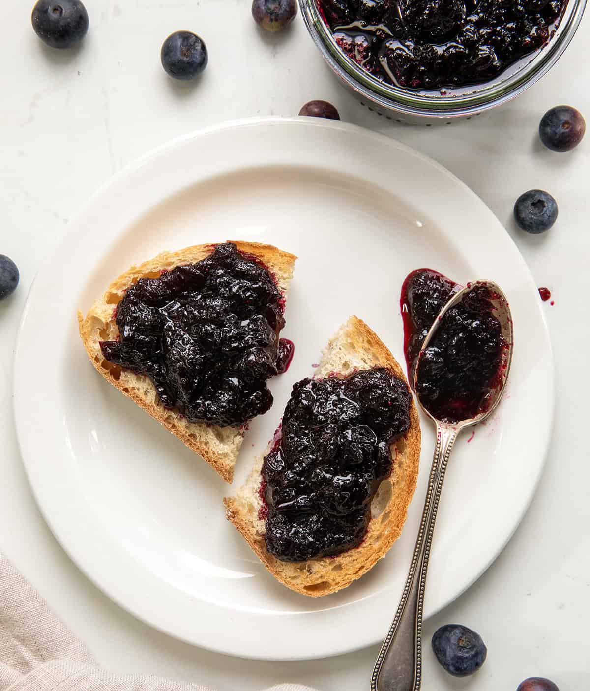 This Bourbon Blueberry Jam is thick, rich, and just sweet enough, with a warm depth of flavor from the bourbon... but does not taste like alcohol at all! Itโs made with pectin, so it sets up nicely and comes together quickly. If you have tried my blueberry jam without pectin (or bourbon) and loved it, I think youโll really enjoy this version, too! Bourbon Blueberry Jam on toast on a white plate on a white table from overhead.